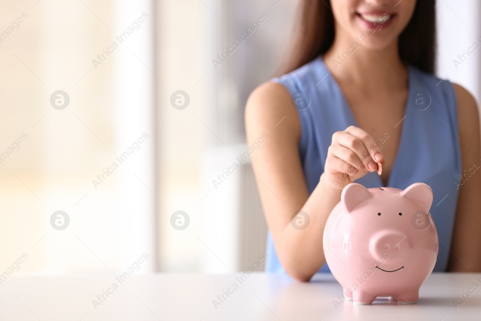 Photo of Woman putting coin into piggy bank at table indoors, closeup. Space for text