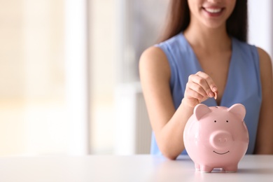 Photo of Woman putting coin into piggy bank at table indoors, closeup. Space for text