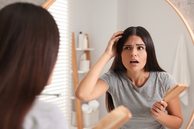 Emotional woman with brush examining her hair and scalp near mirror in bathroom. Dandruff problem