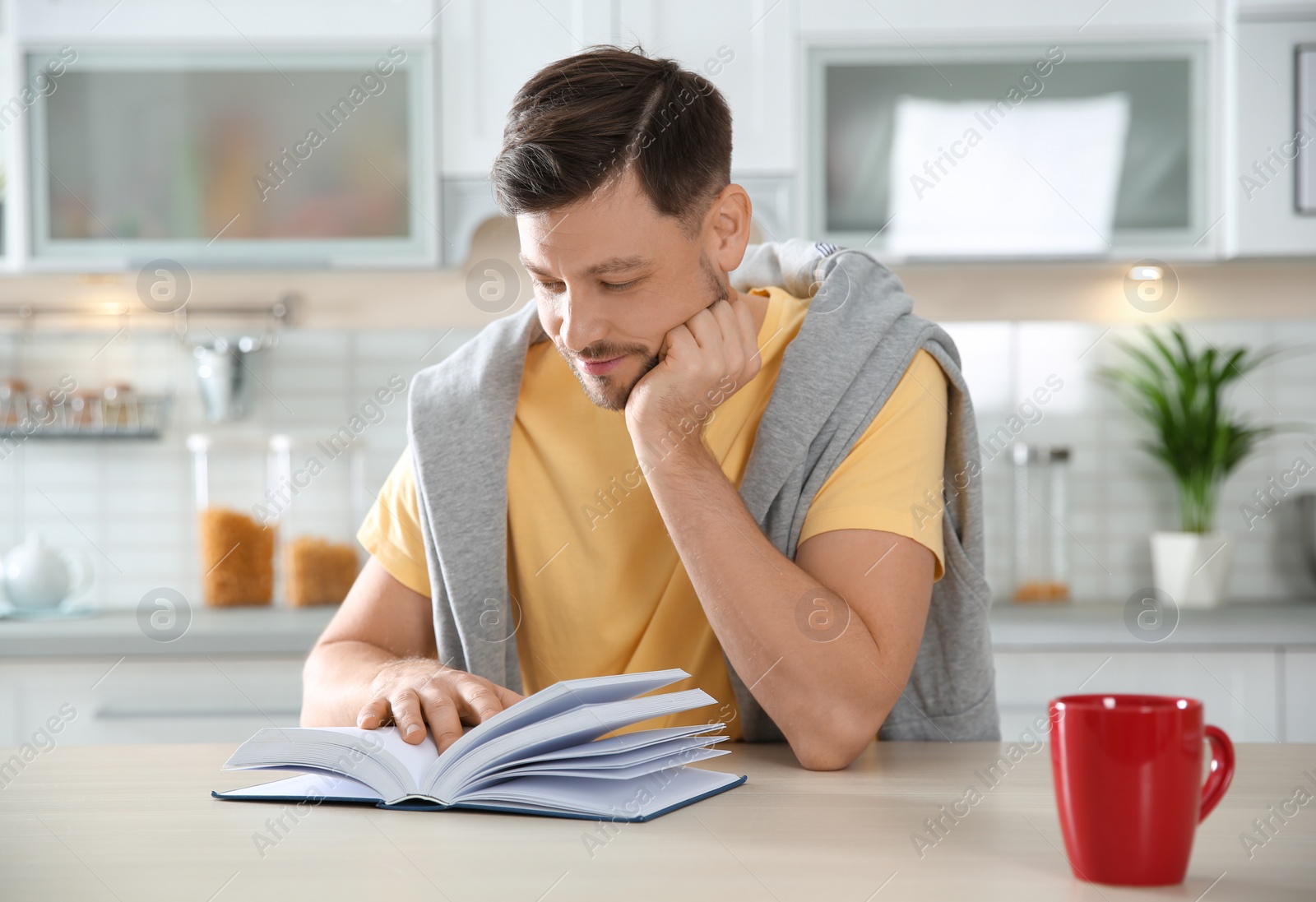 Photo of Handsome man with cup of coffee reading book at table in kitchen