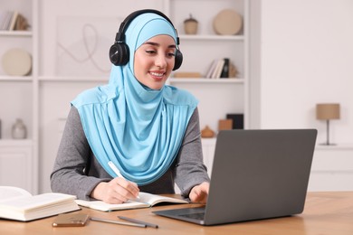 Muslim woman in headphones writing notes near laptop at wooden table in room