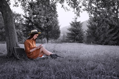 Image of Beautiful young woman drawing with pencil in notepad near tree on grass, color accent effect. State of mindfulness