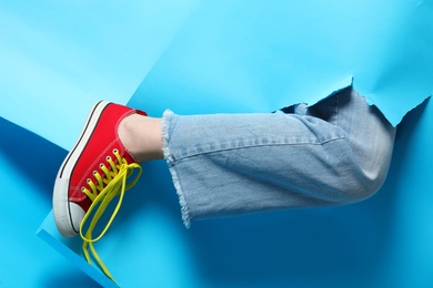 Photo of Woman in stylish shoes with yellow laces showing her leg through torn light blue paper, closeup