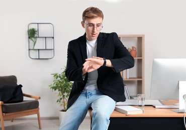 Photo of Young man checking time while sitting on table in office. Being late
