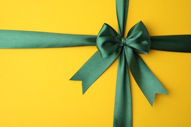 Green satin ribbon with bow on yellow background, top view