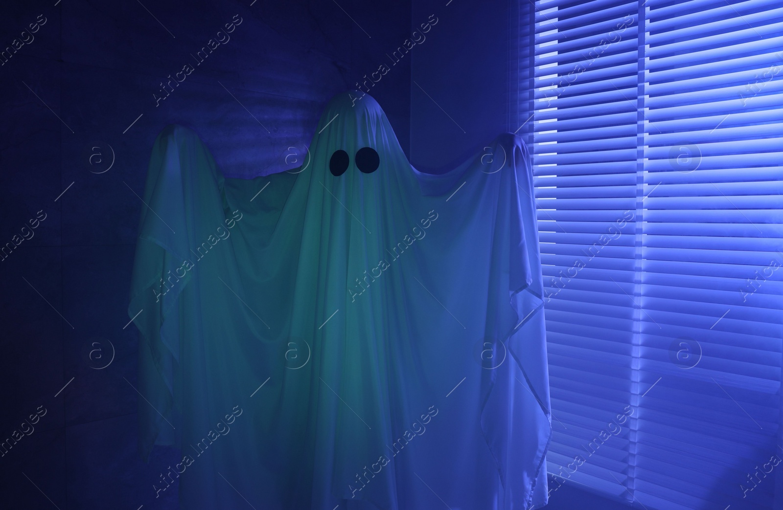 Photo of Creepy ghost. Woman covered with sheet near window in blue light