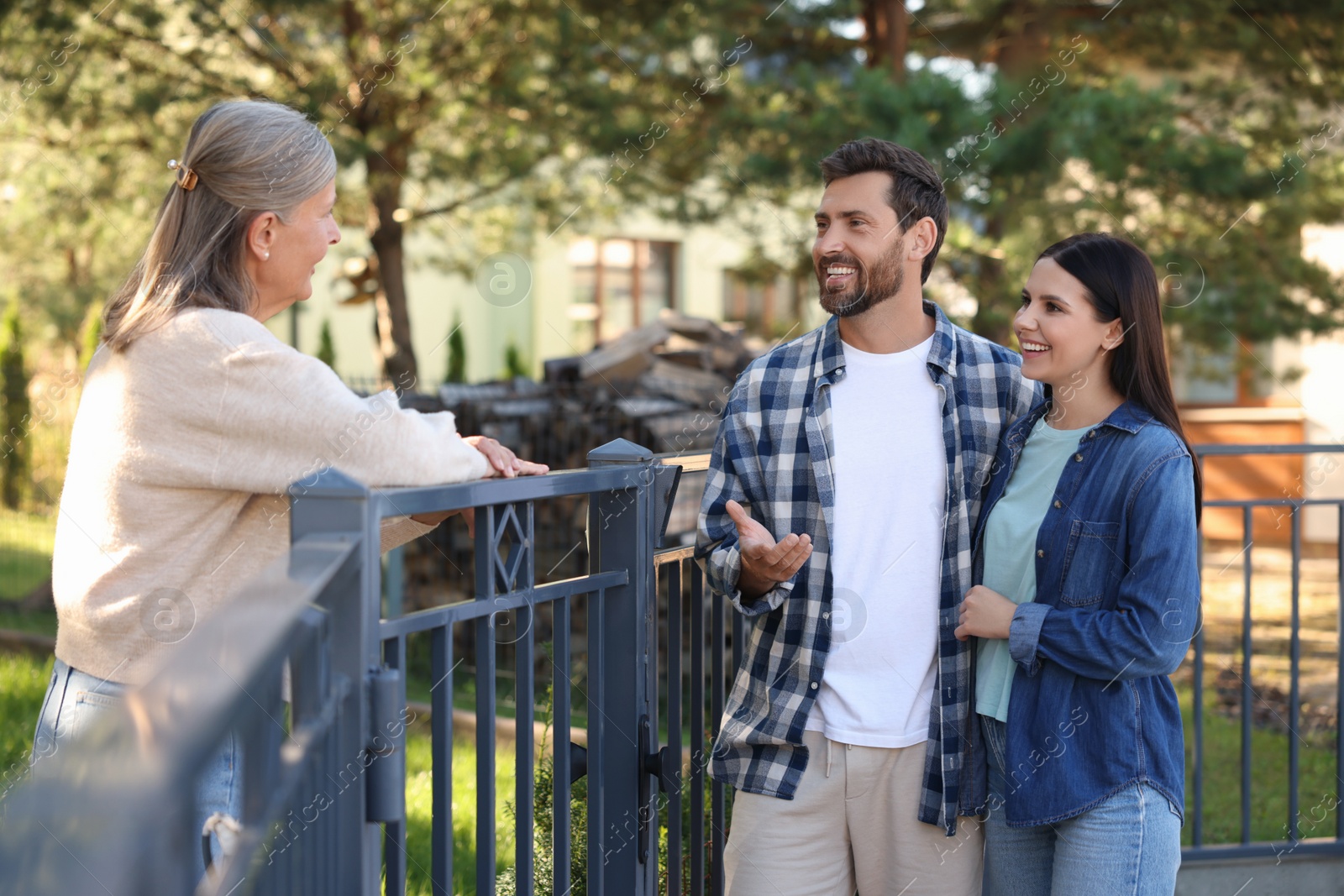 Photo of Friendly relationship with neighbours. Happy young couple talking to senior woman near fence outdoors