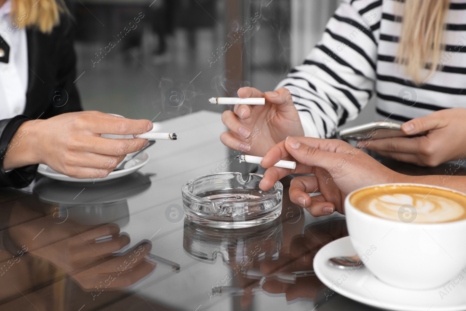 Photo of Women holding cigarettes over glass ashtray at table, closeup