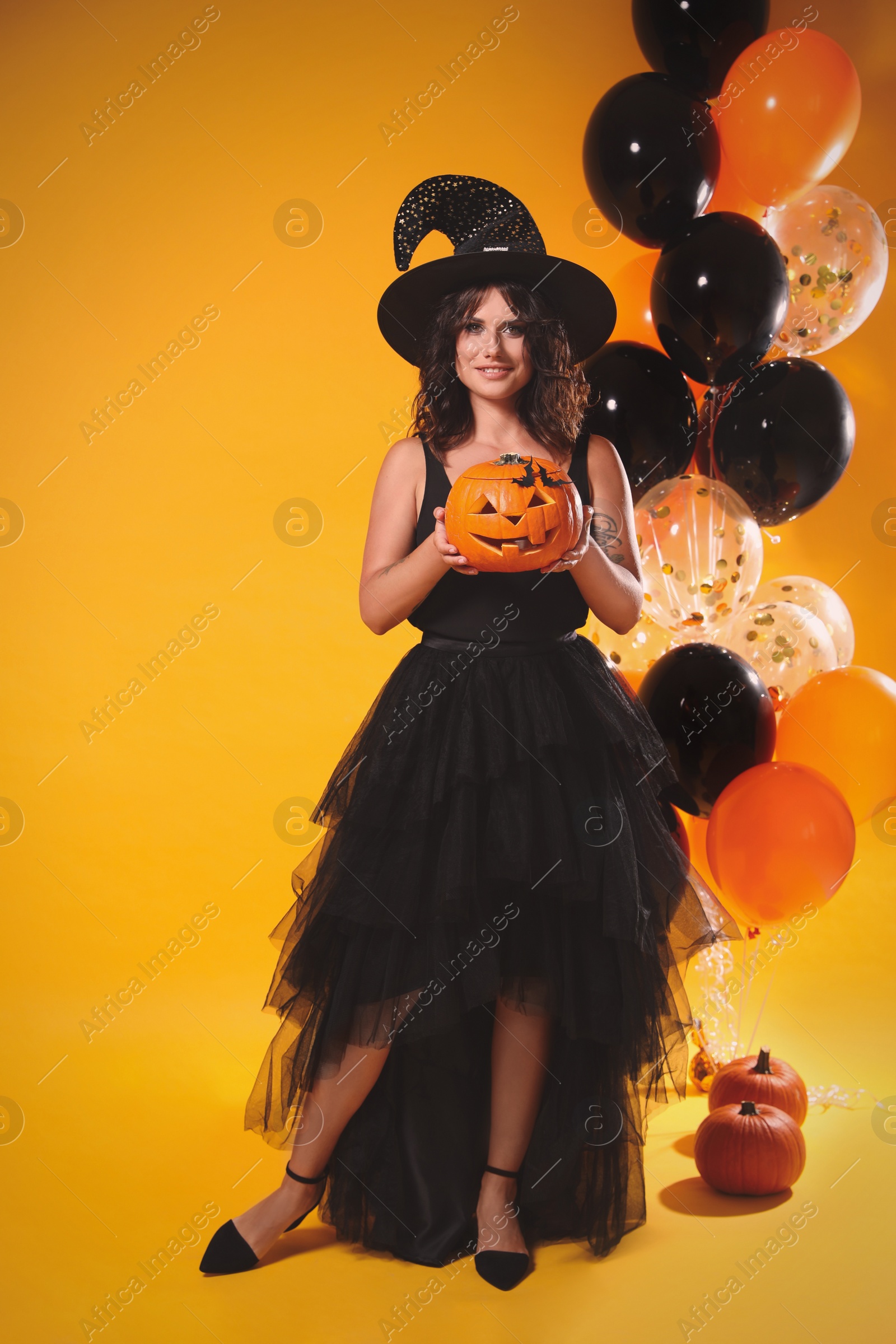 Photo of Beautiful woman in witch costume with balloons and pumpkins on yellow background. Halloween party