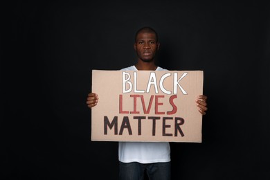 Photo of African American man holding sign with phrase Black Lives Matter on dark background. Racism concept