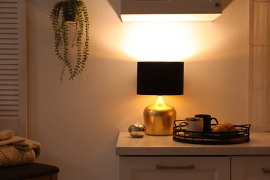 Photo of Stylish lamp, cups and croissant on white cabinet in room. Interior element