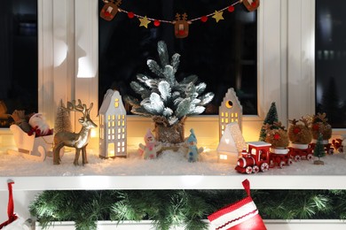Photo of Christmas atmosphere. Beautiful glowing houses, artificial snow and toys on window sill indoors