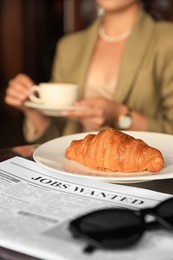 Photo of Woman sitting with cup of hot drink at table, focus on croissant