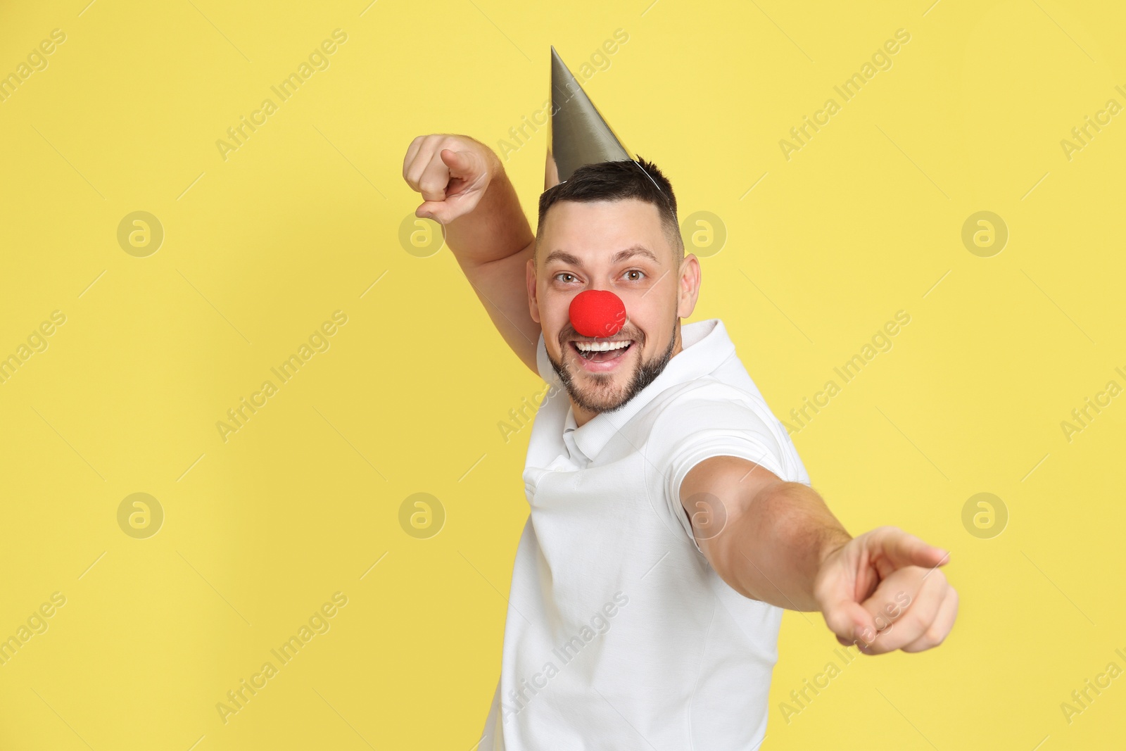 Photo of Funny man with clown nose and party hat on yellow background. April fool's day