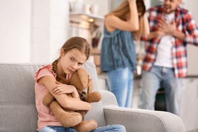 Little unhappy girl sitting on sofa while parents arguing at home