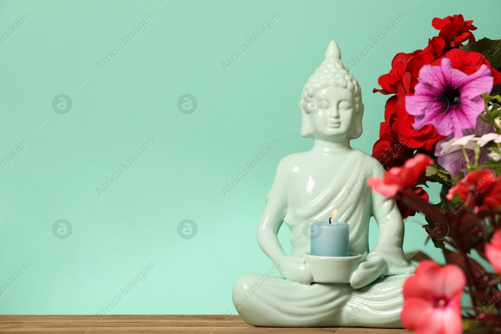 Photo of Buddhism religion. Decorative Buddha statue with burning candle and beautiful flowers on table against turquoise wall, space for text