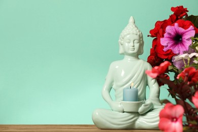 Buddhism religion. Decorative Buddha statue with burning candle and beautiful flowers on table against turquoise wall, space for text