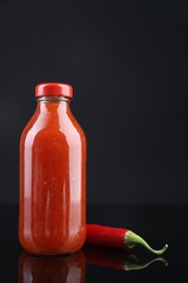 Photo of Spicy chili sauce in bottle and pepper against dark background, space for text