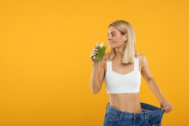 Photo of Slim woman with glass of fresh celery juice wearing big jeans on orange background. Space for text