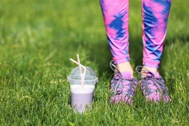 Young woman in sportswear with plastic cup of healthy smoothie on grass outdoors