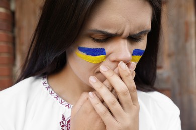 Photo of Sad young woman with drawings of Ukrainian flag on face outdoors, closeup