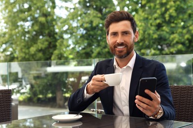 Photo of Handsome bearded man with cup of drink and smartphone at table in cafe