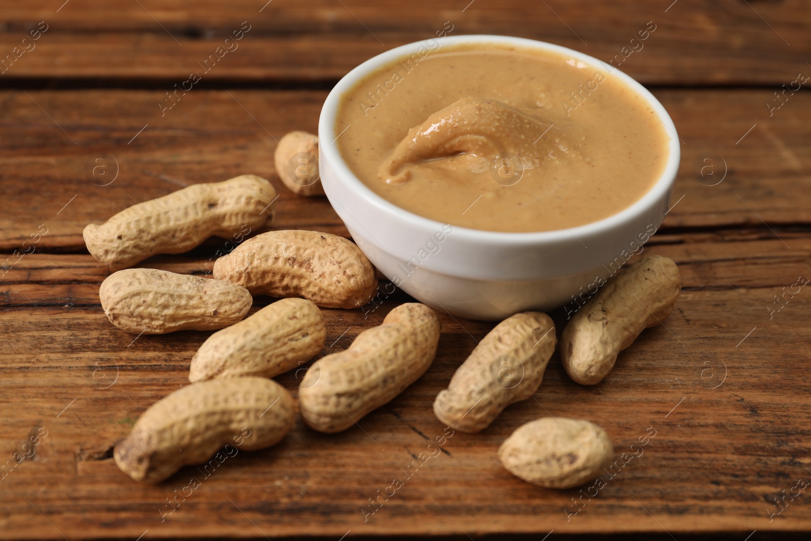 Photo of Delicious nut butter in bowl and peanuts on wooden table, closeup