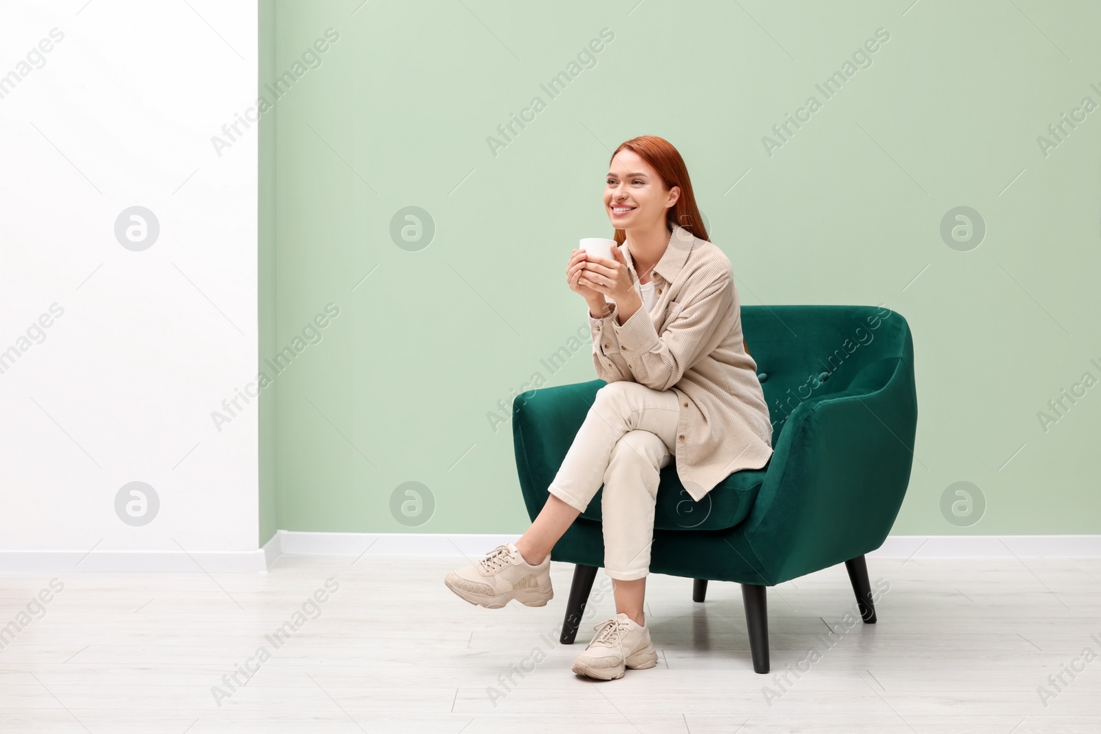 Photo of Happy young woman with cup of drink sitting in armchair indoors. Space for text