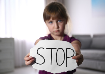 Abused little girl with sign STOP indoors, focus on hands. Domestic violence concept