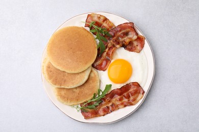 Photo of Plate with tasty pancakes, fried egg, arugula and bacon on light grey textured table, top view