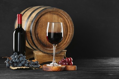 Delicious wine, wooden barrel and fresh grapes on black table, space for text