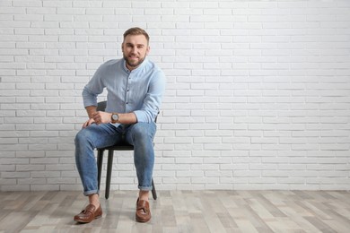 Young man sitting on chair near white brick wall in office, space for text