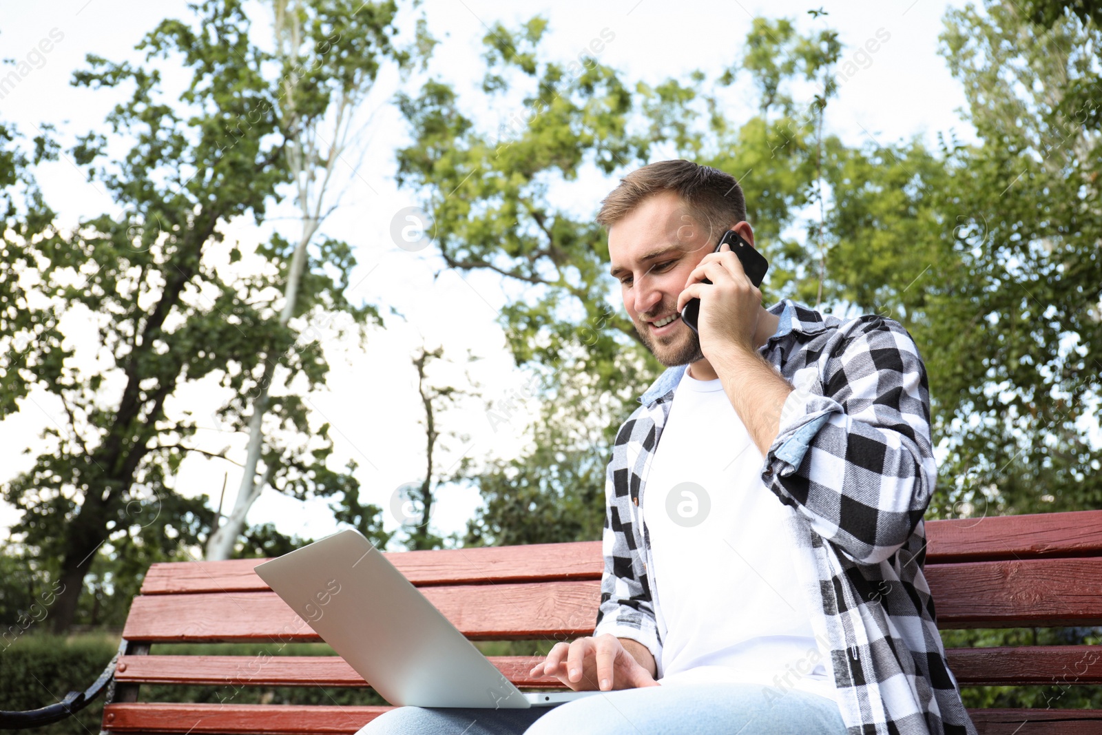 Photo of Young man talking on smartphone while working with laptop in park