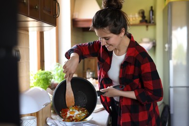 Photo of Young woman putting freshly fried eggs with vegetables onto plate in kitchen