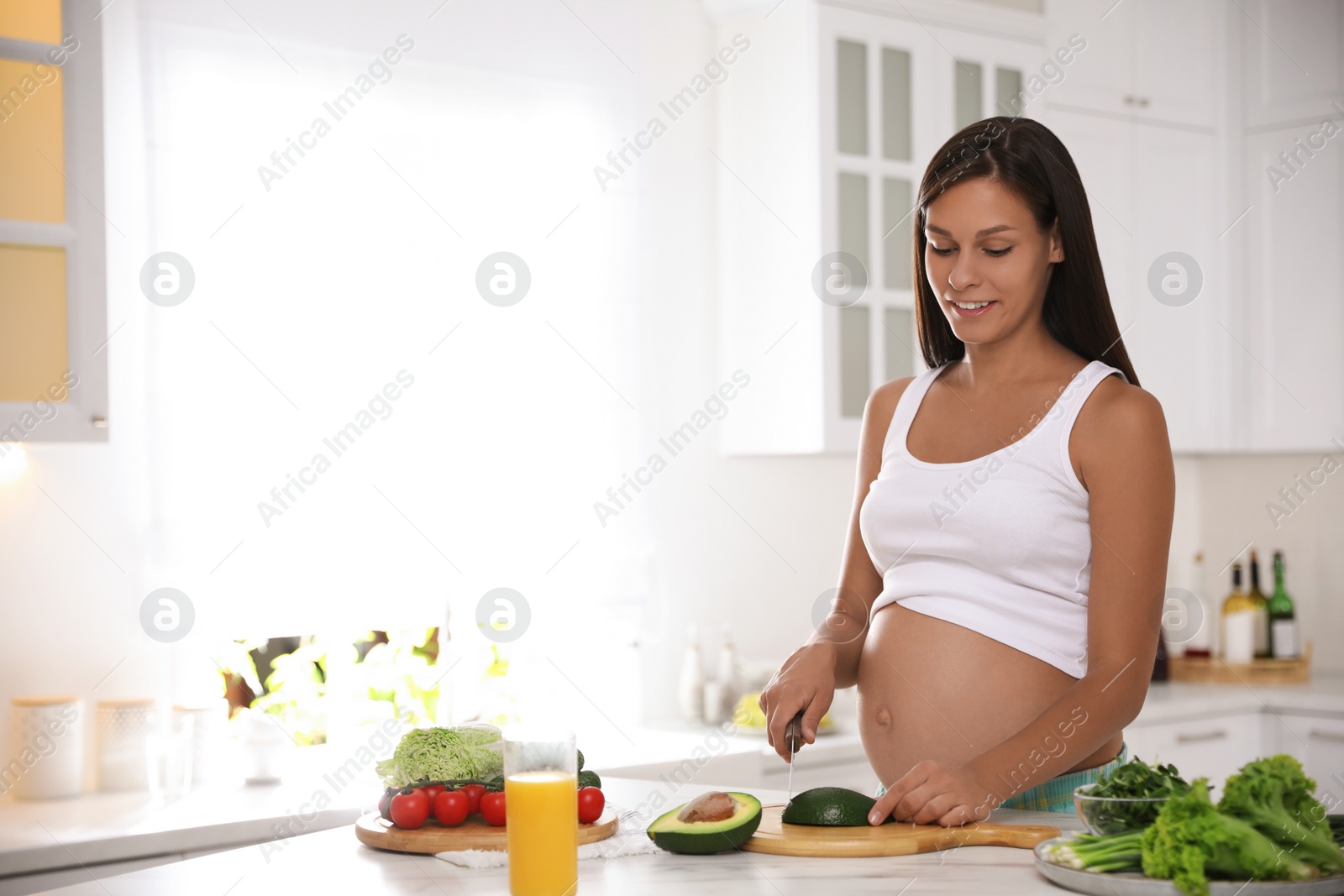 Photo of Young pregnant woman cutting avocado at table in kitchen. Taking care of baby health