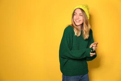 Happy young woman wearing warm sweater and knitted hat on yellow background. Space for text