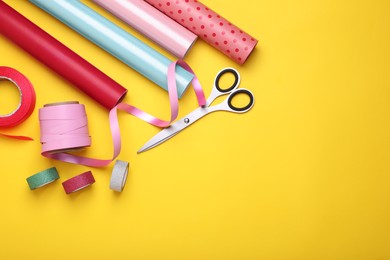 Rolls of colorful wrapping papers, scissors and ribbons on yellow background, flat lay. Space for text