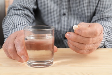 Senior man holding pill and glass of water at table, closeup