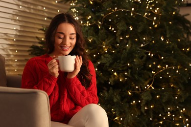 Photo of Beautiful woman with cup of tasty drink near Christmas tree indoors