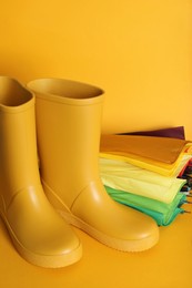 Photo of Pair of bright rubber boots near umbrella on pale orange background