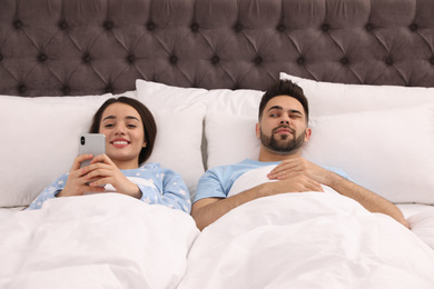 Distrustful young man peering into girlfriend's smartphone in bed at home