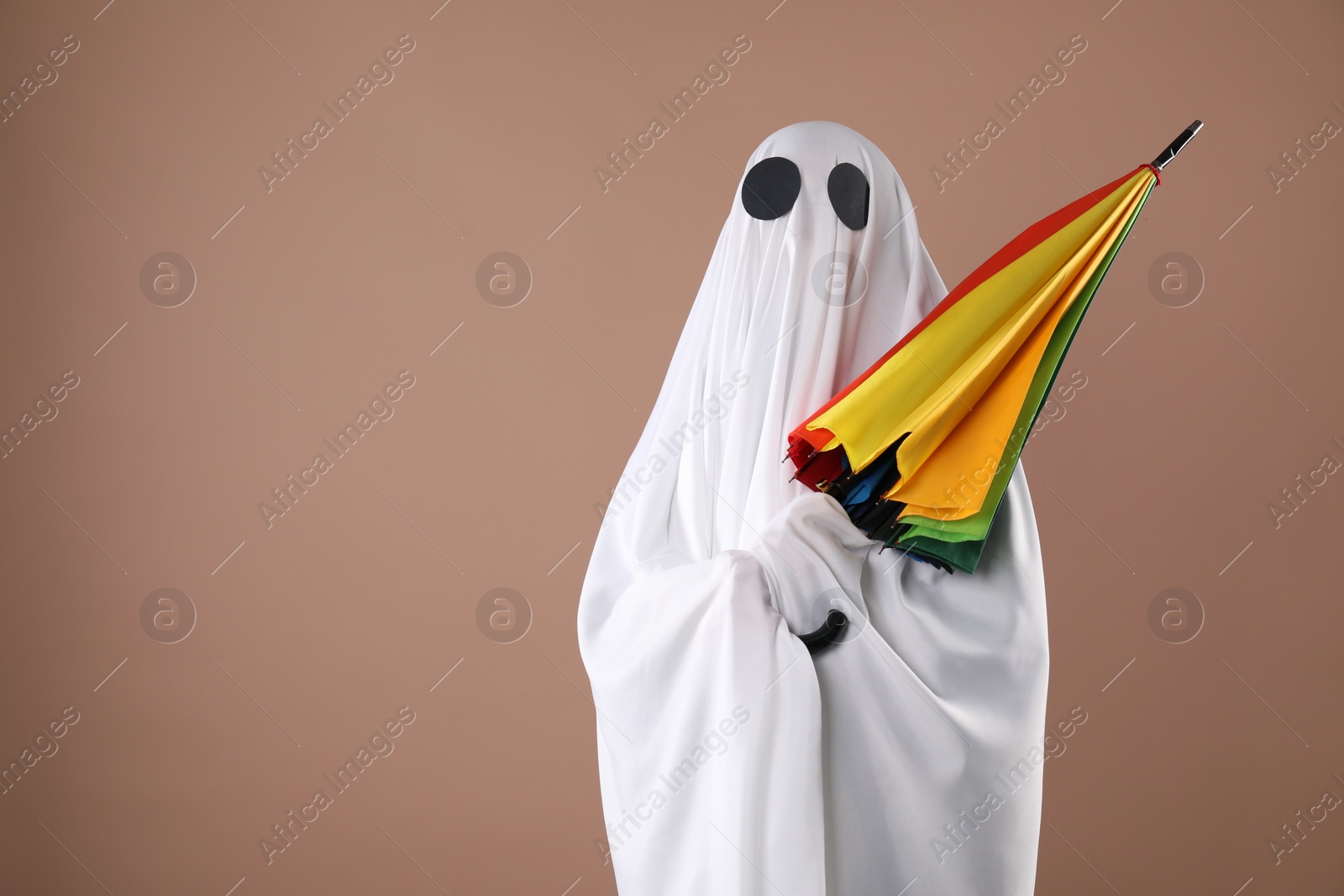 Photo of Person in ghost costume with rainbow umbrella on dark beige background, space for text