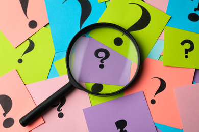 Photo of Magnifier glass on different colorful paper cards with question marks, top view