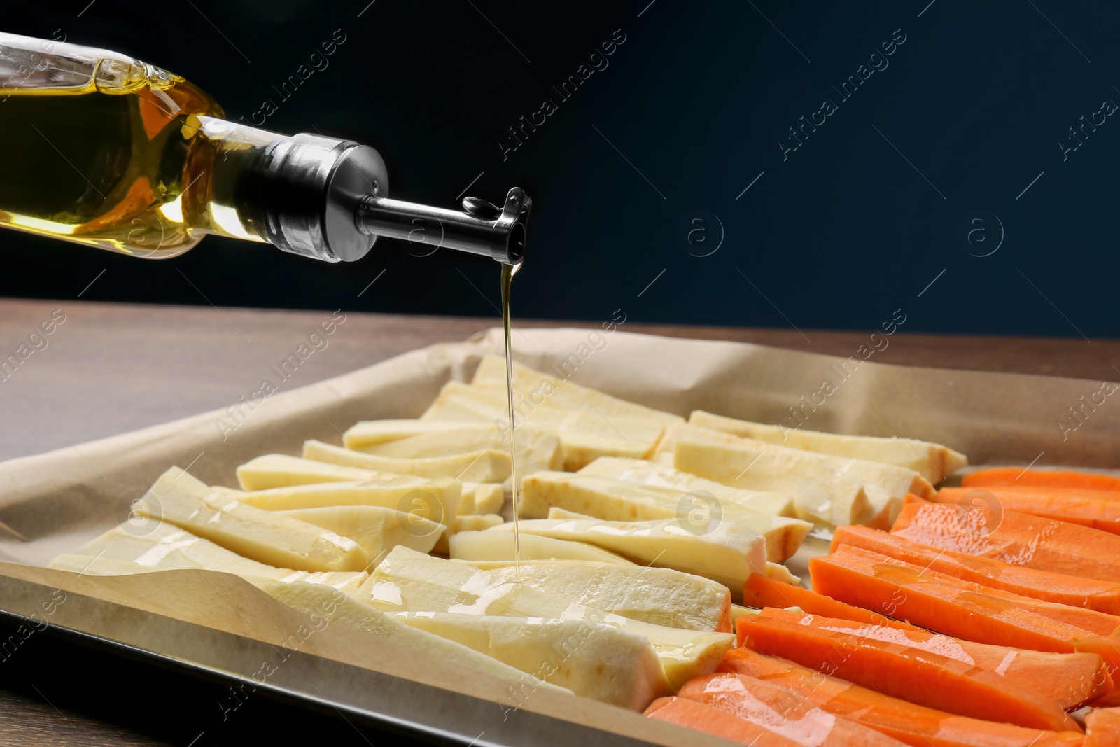 Photo of Pouring oil onto baking tray with parsnips and carrots against blue background, closeup