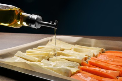 Pouring oil onto baking tray with parsnips and carrots against blue background, closeup