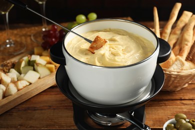 Dipping piece of bread into fondue pot with melted cheese at wooden table, closeup