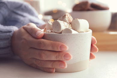 Photo of Woman holding cup of delicious hot cocoa with marshmallows at white table indoors, closeup. Winter drink
