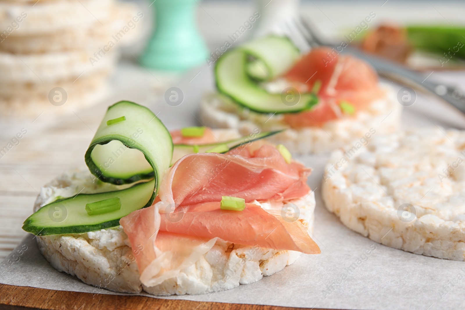 Photo of Puffed rice cakes with prosciutto and cucumber on wooden board, closeup