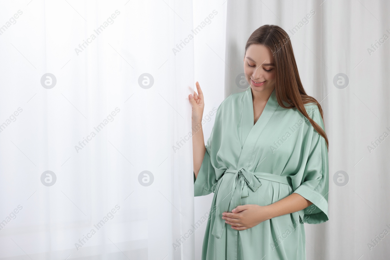 Photo of Beautiful pregnant woman in green dressing gown near window indoors, space for text