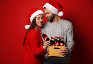 Photo of MYKOLAIV, UKRAINE - JANUARY 27, 2021: Young couple in Christmas hats holding crate with bottles of Coca-Cola on red background